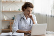 © fizkes - Concerned young female Caucasian doctor in white medical uniform sit at desk in hospital work on laptop, focused woman nurse or GP busy fill anamnesis on computer gadget, medicine, technology concept