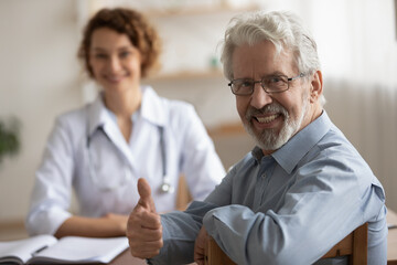 Wall Mural - Smiling mature male patient sit at consultation with doctor in clinic show thumb up recommend good quality medical company service, portrait of happy senior man client give recommendation to hospital