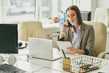 Wall Mural - Young pretty brunette office worker with protective mask on chin using tablet