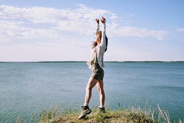 Wall Mural - Young ecstatic woman with her arms raised standing on riverbank by water