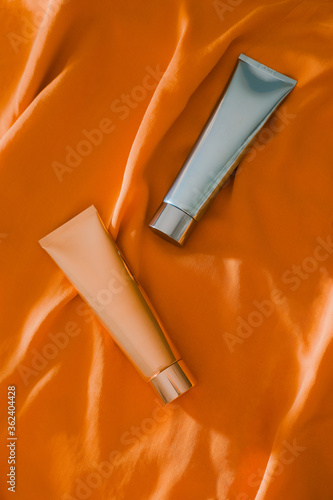 Shiny light blue and golden cosmetic tubes on sunny orange fabric background Poster Mural XXL