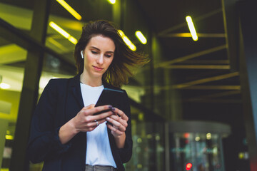 Wall Mural - Young female student standing near neon yellow light in night city and listening to music in wireless earphones on modern smartphone. elegant woman watching media video on cellphone device