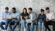 © fizkes - Six multi racial millennial girls and guys sit in line on chairs against grey wall using diverse devices, busy students discuss tasks, employees wait seminar in corridor having fun in internet concept