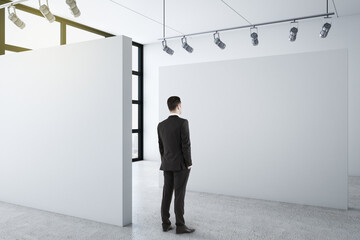 Wall Mural - Businessman standing in clean gallery interior and looking on blank wall.