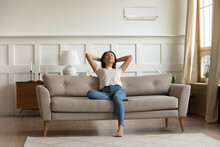 Happy Young Calm Woman Relaxing On Sofa, Breathing Fresh Air, Satisfied With Comfortable Indoors Temperature. Enjoying Spending Hot Summer Time In Cooled Living Room, Front Full Length View.