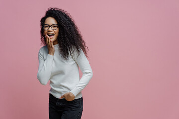 Wall Mural - Positive African American woman laughs at something funny, covers opened mouth with palm, cannot control her emotions, keeps hand in pocket of jeans, wears white poloneck, isolated on pink background