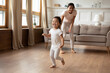 Full length overjoyed little preschool cute vietnamese ethnic baby girl running barefoot on warm floor, having fun playing with energetic young asian mother or nanny, leisure daycare activity.