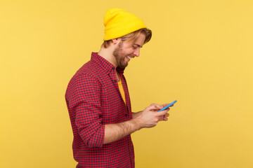 Wall Mural - side view happy trendy guy in hipster beanie hat and shirt using cell phone, texting or reading mess
