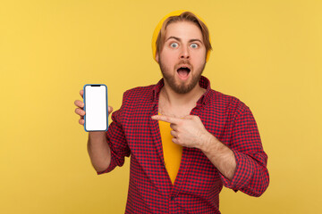 Wall Mural - shocked trendy hipster guy in beanie hat and checkered shirt pointing mobile phone empty display, lo