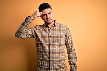 Young handsome man wearing casual shirt standing over isolated yellow background pointing unhappy to pimple on forehead, ugly infection of blackhead. Acne and skin problem