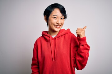 Young beautiful asian girl wearing casual sweatshirt with hoodie over white background smiling with happy face looking and pointing to the side with thumb up.