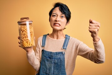 Young beautiful asian girl holding jar with italian dry pasta macaroni over yellow background annoyed and frustrated shouting with anger, crazy and yelling with raised hand, anger concept