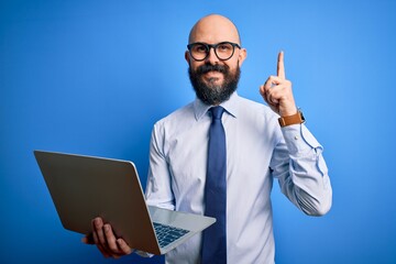 Handsome bald business man with beard working using laptop over blue background surprised with an idea or question pointing finger with happy face, number one