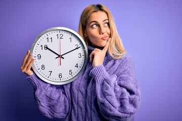 Poster - Young beautiful blonde woman doing countdown holding big clock over purple background serious face thinking about question, very confused idea