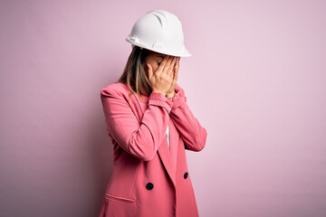 Poster - Young beautiful brunette architect woman wearing safety helmet over pink background with sad expression covering face with hands while crying. Depression concept.