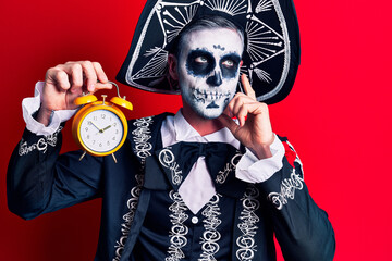 Canvas Print - Young man wearing mexican day of the dead costume holding alarm clock serious face thinking about question with hand on chin, thoughtful about confusing idea