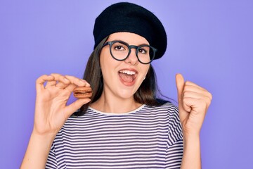 Poster - Young beautiful girl wearing glasses and fashion beret holding french sweet pastry macaron screaming proud and celebrating victory and success very excited, cheering emotion