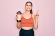 Young beautiful fitness woman wearing sport excersie clothes drinking protein shake surprised with an idea or question pointing finger with happy face, number one