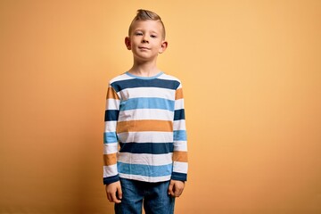 Wall Mural - Young little caucasian kid with blue eyes wearing colorful striped shirt over yellow background Relaxed with serious expression on face. Simple and natural looking at the camera.