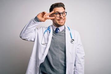 Wall Mural - Young doctor man with blue eyes wearing medical coat and stethoscope over isolated background Doing peace symbol with fingers over face, smiling cheerful showing victory