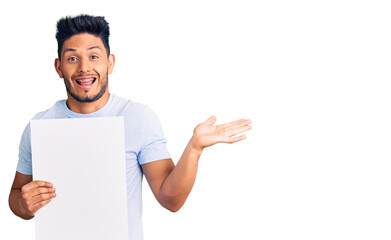 Handsome latin american young man holding cardboard banner with blank space celebrating victory with happy smile and winner expression with raised hands