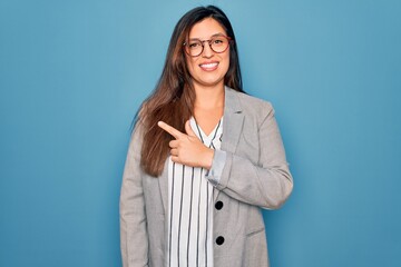 Sticker - Young hispanic business woman wearing glasses standing over blue isolated background cheerful with a smile on face pointing with hand and finger up to the side with happy and natural expression