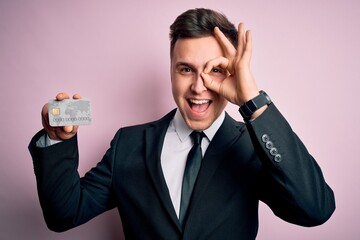 Wall Mural - Young handsome caucasian business man holding finance credit card over pink background with happy face smiling doing ok sign with hand on eye looking through fingers