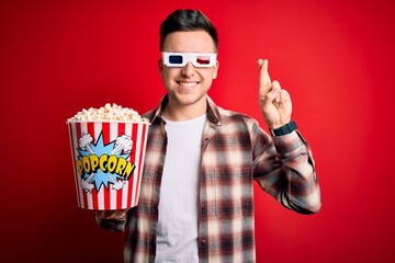 Wall Mural - Young handsome caucasian man wearing 3d movie glasses and eating popcorn gesturing finger crossed smiling with hope and eyes closed. Luck and superstitious concept.