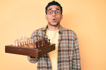 Canvas Print - Young handsome hispanic man holding chess board wearing glasses scared and amazed with open mouth for surprise, disbelief face