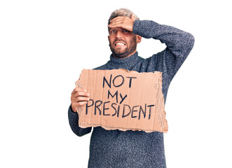 Young handsome blond man holding not my president cardboard banner stressed and frustrated with hand on head, surprised and angry face