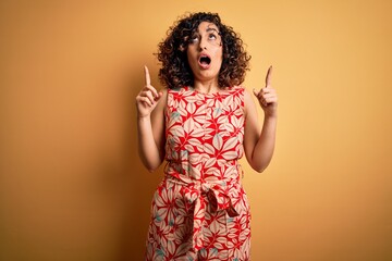 Poster - Young beautiful curly arab woman on vacation wearing summer floral dress and sunglasses amazed and surprised looking up and pointing with fingers and raised arms.