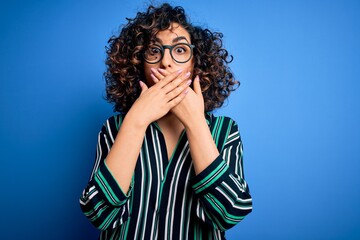 Poster - Young beautiful curly arab woman wearing striped shirt and glasses over blue background shocked covering mouth with hands for mistake. Secret concept.