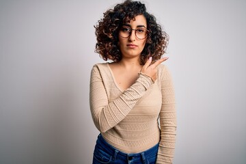 Poster - Young beautiful curly arab woman wearing casual t-shirt and glasses over white background Pointing with hand finger to the side showing advertisement, serious and calm face