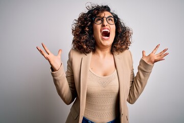 Poster - Young curly arab business woman wearing elegant jacket and glasses over white background crazy and mad shouting and yelling with aggressive expression and arms raised. Frustration concept.