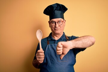 Wall Mural - Middle age cooker man wearing apron and hat holding wooden spoon over yellow background with angry face, negative sign showing dislike with thumbs down, rejection concept