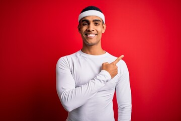 Wall Mural - Young handsome african american sportsman wearing sportswear over red background cheerful with a smile on face pointing with hand and finger up to the side with happy and natural expression