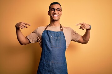 Wall Mural - Young handsome african american shopkeeper man wearing apron over yellow background looking confident with smile on face, pointing oneself with fingers proud and happy.