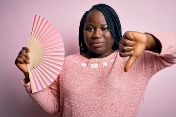 Wall Mural - Young african american plus size woman with braids using hand fan over pink background with angry face, negative sign showing dislike with thumbs down, rejection concept