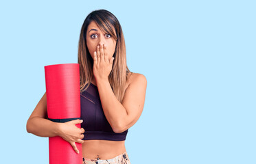 Young beautiful woman wearing sportswear holding yoga mat covering mouth with hand, shocked and afraid for mistake. surprised expression