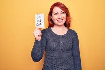 Wall Mural - Young beautiful redhead woman asking for equality holding paper with we are equal message looking positive and happy standing and smiling with a confident smile showing teeth