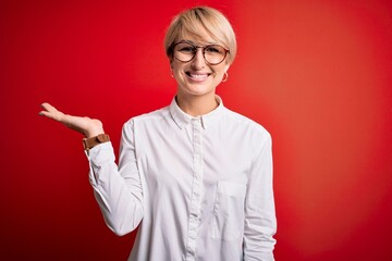 Sticker - Young blonde business woman with short hair wearing glasses over red background smiling cheerful presenting and pointing with palm of hand looking at the camera.