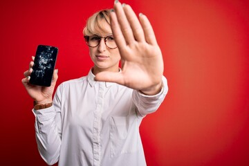 Poster - Young blonde woman with short hair holding broken smartphone over red background with open hand doing stop sign with serious and confident expression, defense gesture