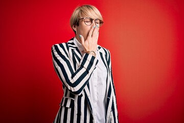 Poster - Blonde business woman with short hair wearing glasses and striped jacket over red background bored yawning tired covering mouth with hand. Restless and sleepiness.