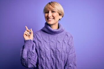 Wall Mural - Young blonde woman with short hair wearing winter turtleneck sweater over purple background with a big smile on face, pointing with hand finger to the side looking at the camera.