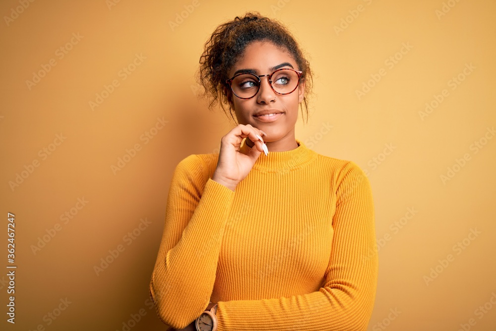 Young beautiful african american girl wearing sweater and glasses over yellow background with hand on chin thinking about question, pensive expression. Smiling and thoughtful face. Doubt concept.