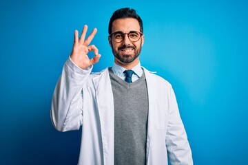 Sticker - Young handsome doctor man with beard wearing coat and glasses over blue background smiling positive doing ok sign with hand and fingers. Successful expression.