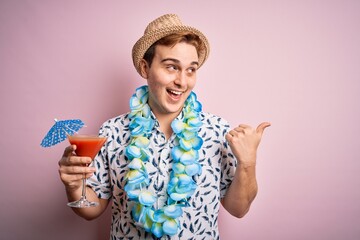 Wall Mural - Young redhead tourist man on vacation wearing hat and hawaiian lei drinking cocktail pointing thumb up to the side smiling happy with open mouth