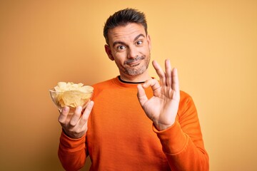 Young handsome man holding bowl with potatoes chips over isolated yellow background doing ok sign with fingers, excellent symbol