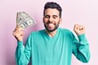 © Krakenimages.com - Young handsome man with beard holding bunch of dollar screaming proud, celebrating victory and success very excited with raised arm