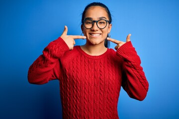 Sticker - Young beautiful asian woman wearing casual sweater and glasses over blue background smiling cheerful showing and pointing with fingers teeth and mouth. Dental health concept.